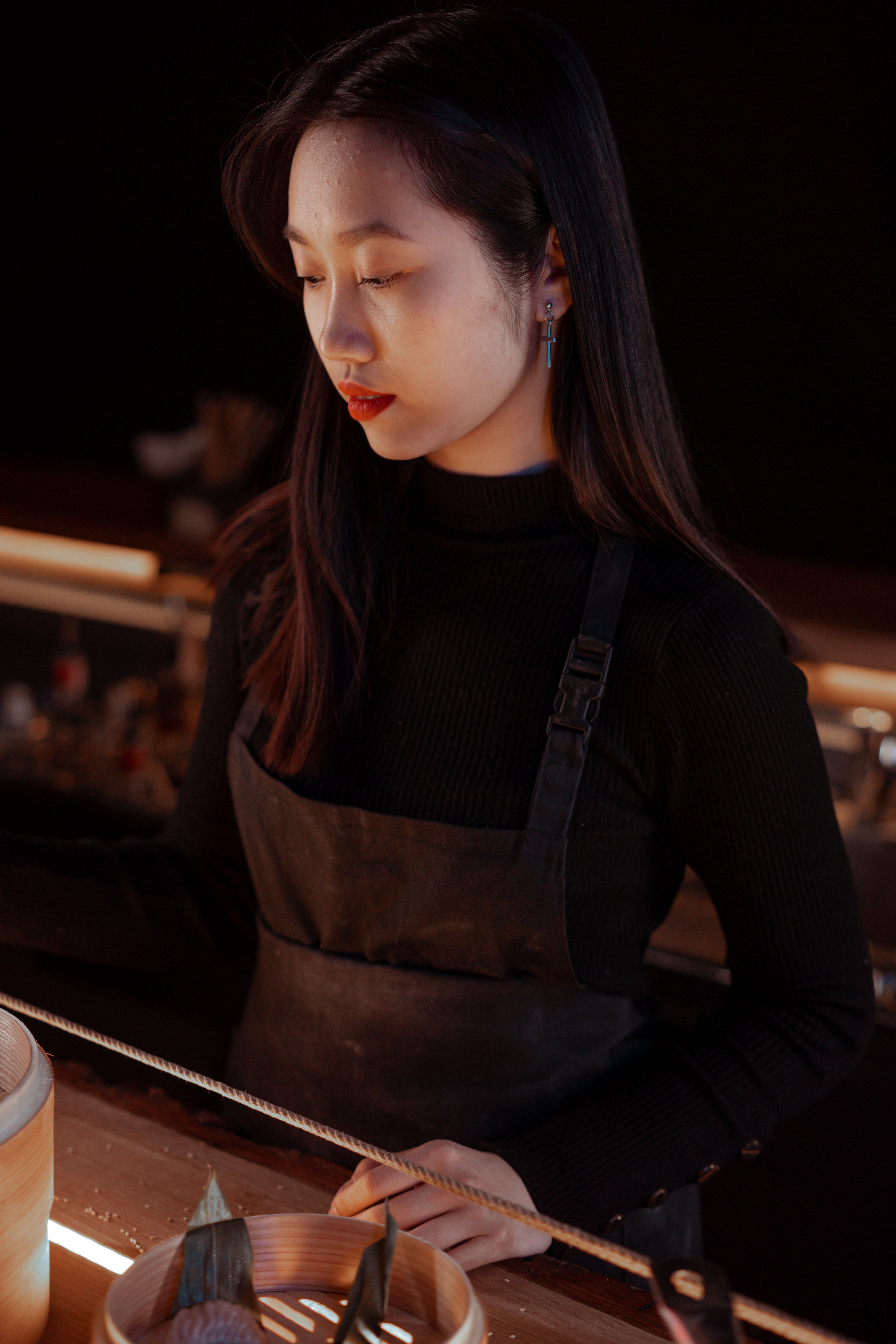 Portrait of a woman with long hair and red lipstick wearing a black apron in a dimly lit setting.