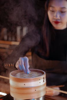 An Asian woman prepares food using a bamboo steamer in a kitchen setting, surrounded by steam.