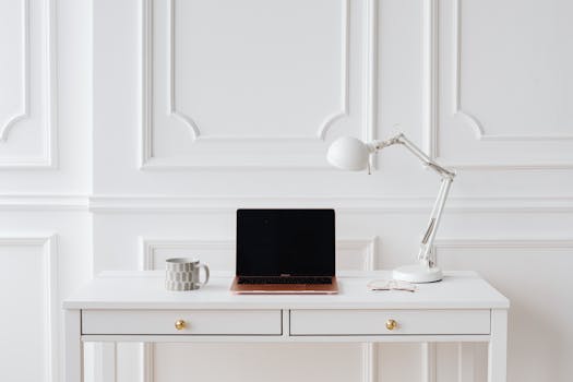Elegant minimalist workspace with laptop and desk lamp on a white desk in a bright room.