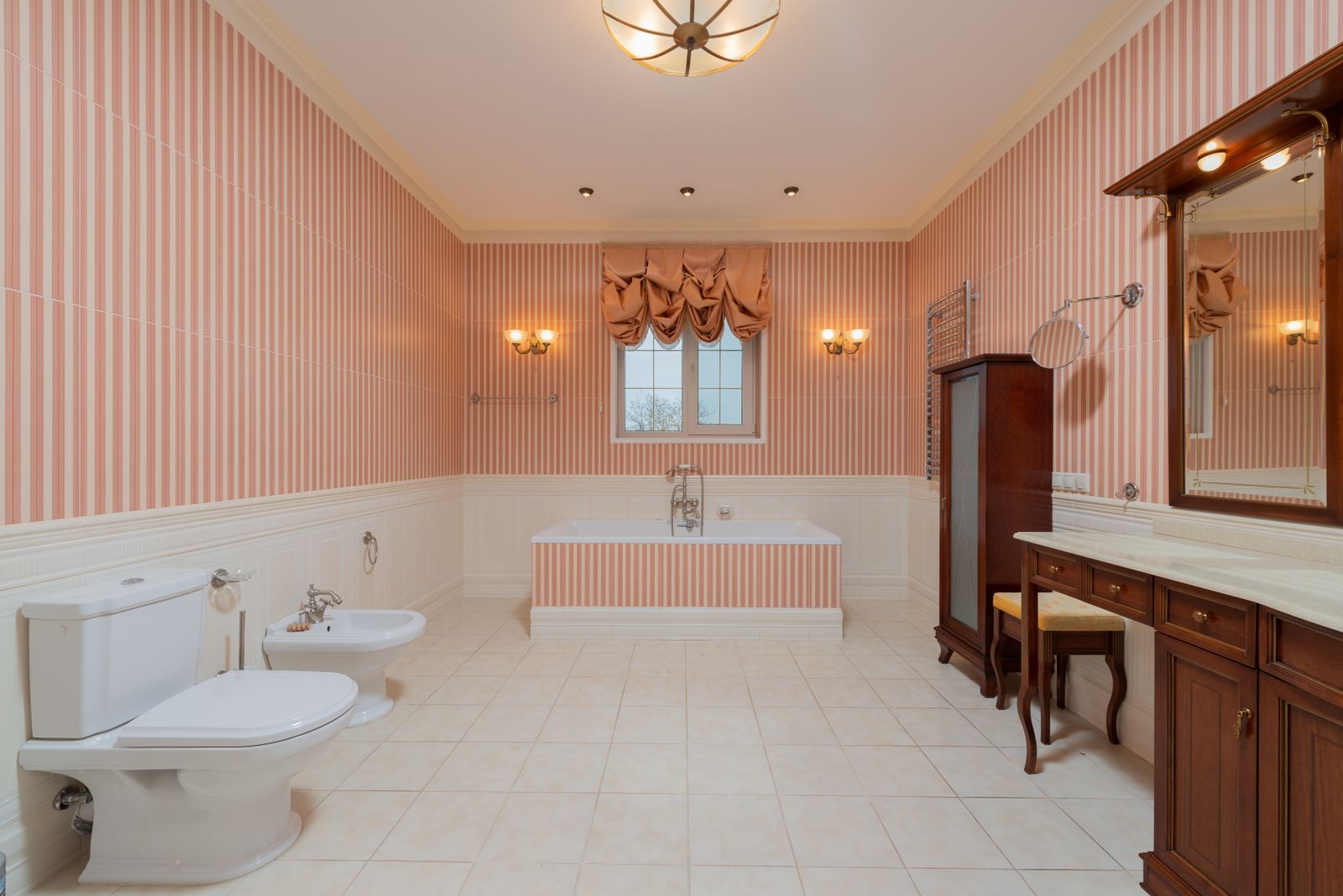 Pink striped wallpaper above white lower-wall paneling, viewed toward a bathtub and window.