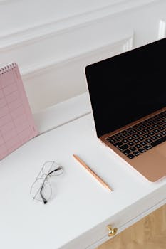 A clean, minimalist desk with a laptop, glasses, pen, and pink calendar.