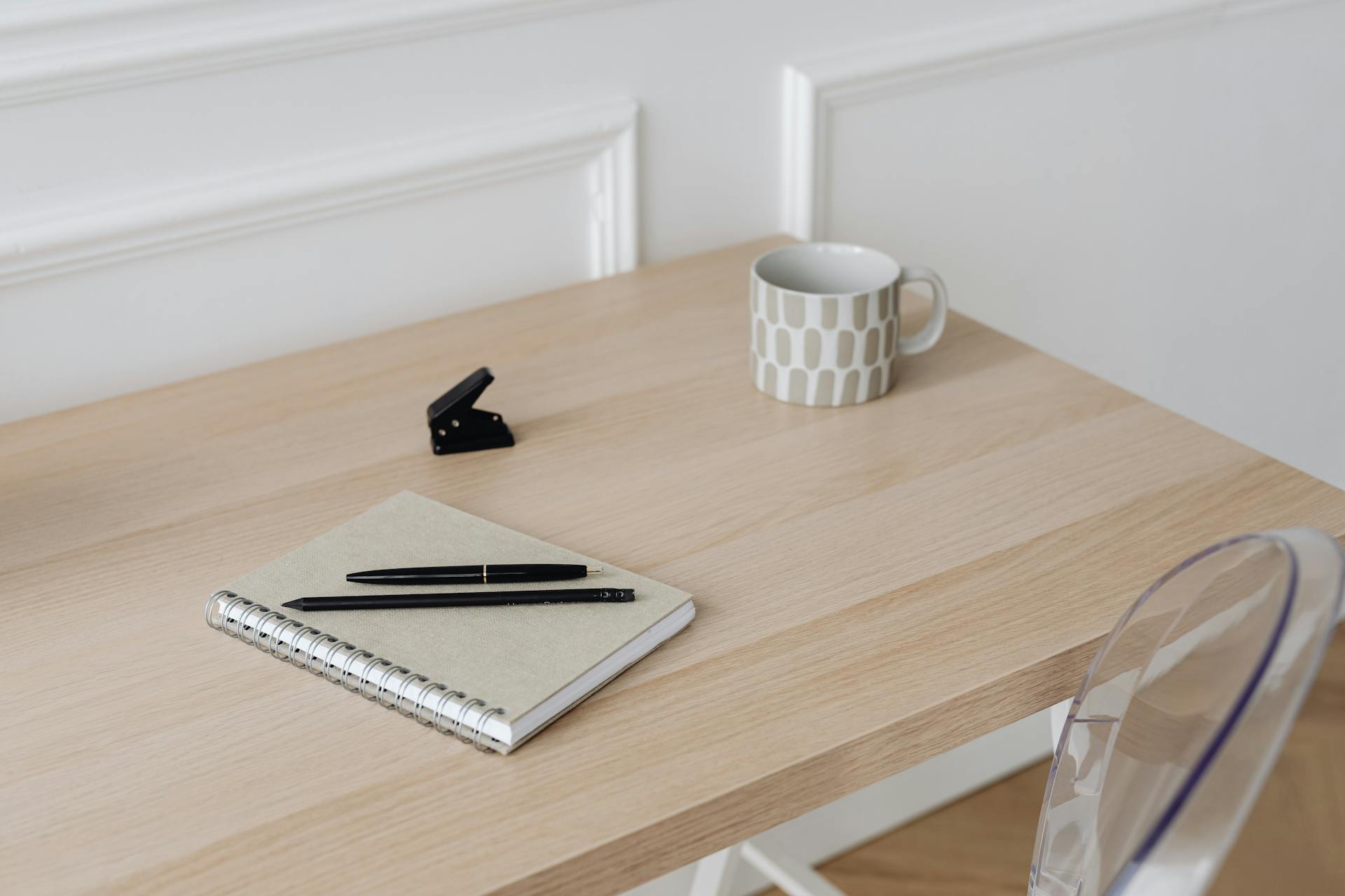 A minimalist desk setup featuring a notebook, pens, mug, and stapler on a wooden table.