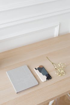 A minimalist flat lay featuring a smartphone, notebook, and delicate flowers on a wooden table.