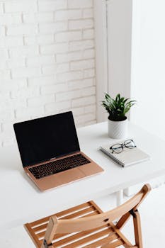 A simple and organized workspace featuring a laptop, notebook, eyeglasses, and a plant on a white desk.