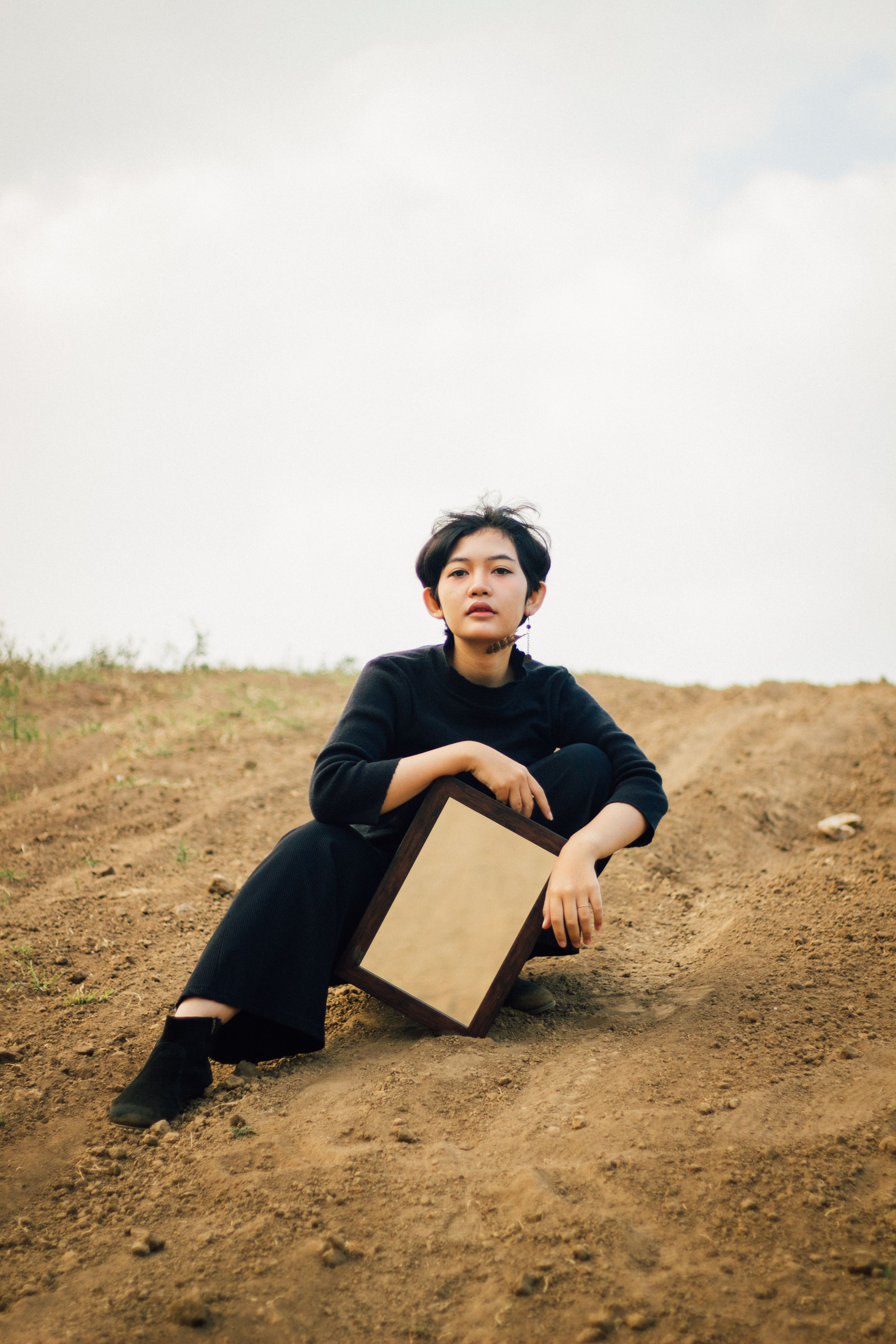 A woman sits outdoors on a hill holding a mirror, evoking introspection.