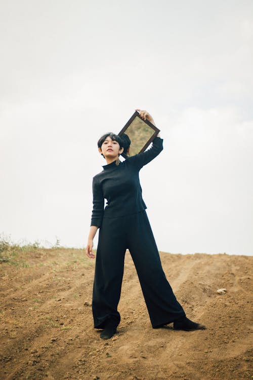 A Low Angle Shot of a Woman in Black Turtleneck Sweater and Pants Standing while Holding a Wooden Frame