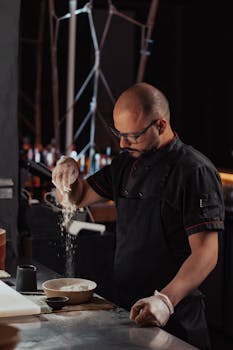 A focused chef sprinkles flour over a dish in a dimly lit restaurant kitchen.