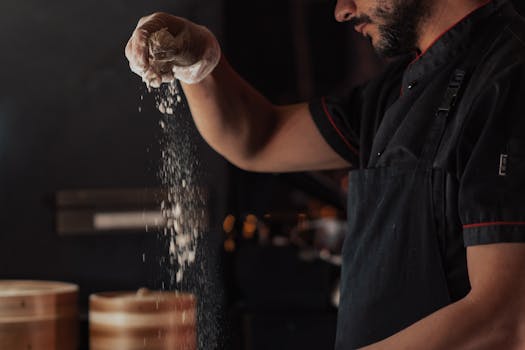 A chef wearing a black apron sprinkles flour onto a surface in a dimly lit kitchen, showcasing food preparation.