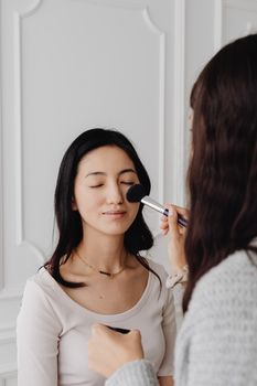A woman with eyes closed gets a makeup brush applied to her face. Captured indoors.