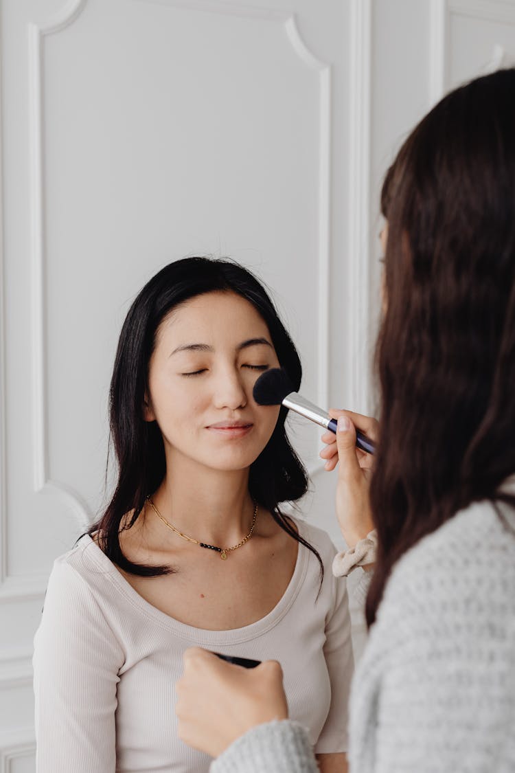 A Person Applying A Makeup On A Woman In White Shirt With Her Eyes Closed