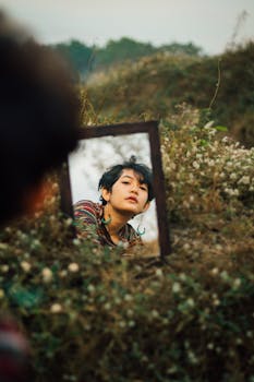 An artistic reflection of a young woman in a mirror surrounded by wildflowers outdoors.