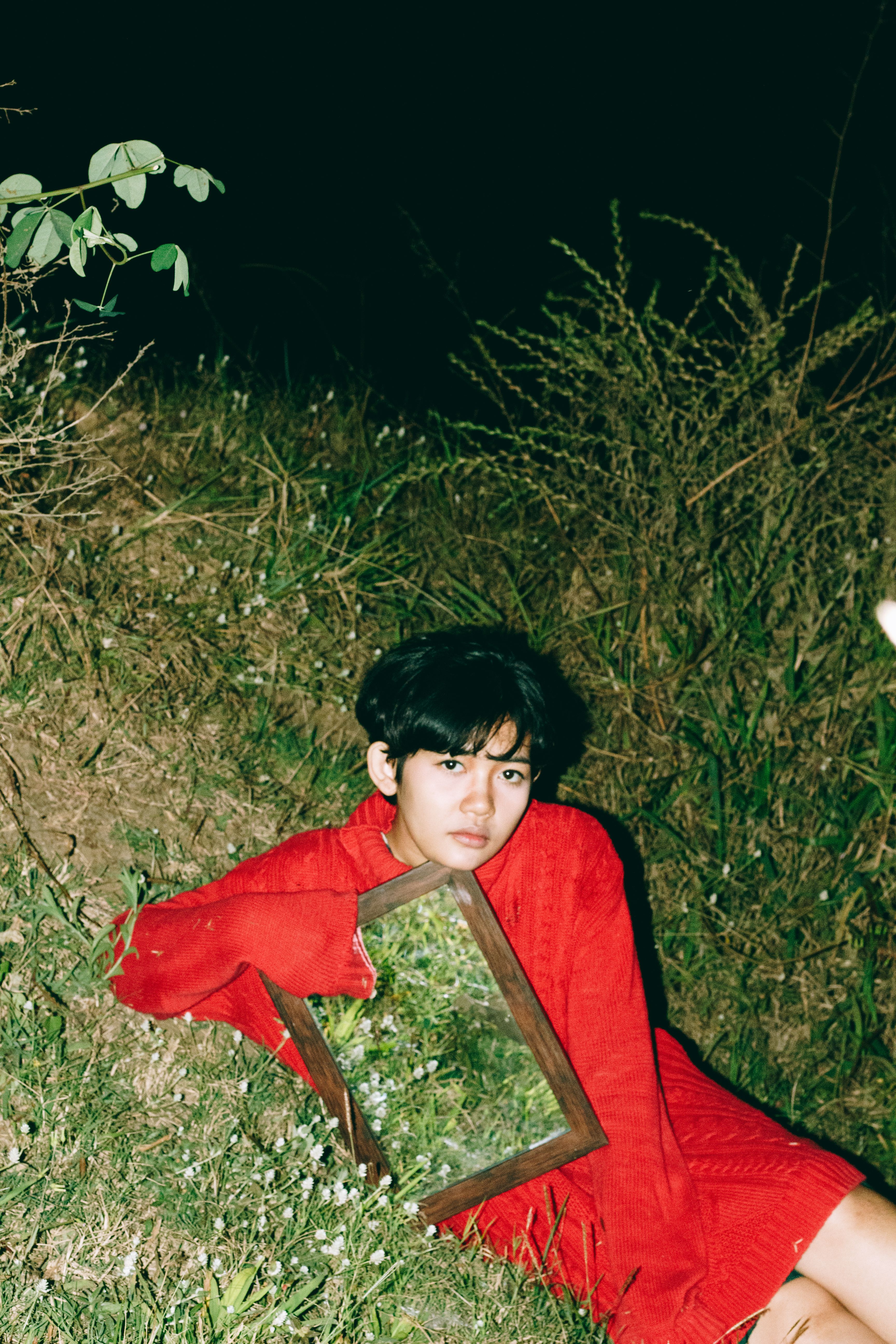 A young woman in a red sweater sitting outside at night holding a mirror reflecting greenery.