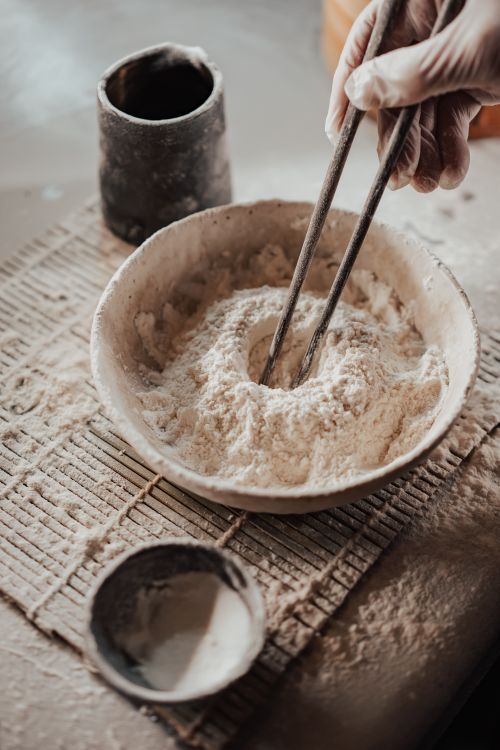 Free A hand stirs flour in a rustic bowl using chopsticks, creating a textured culinary moment. Stock Photo