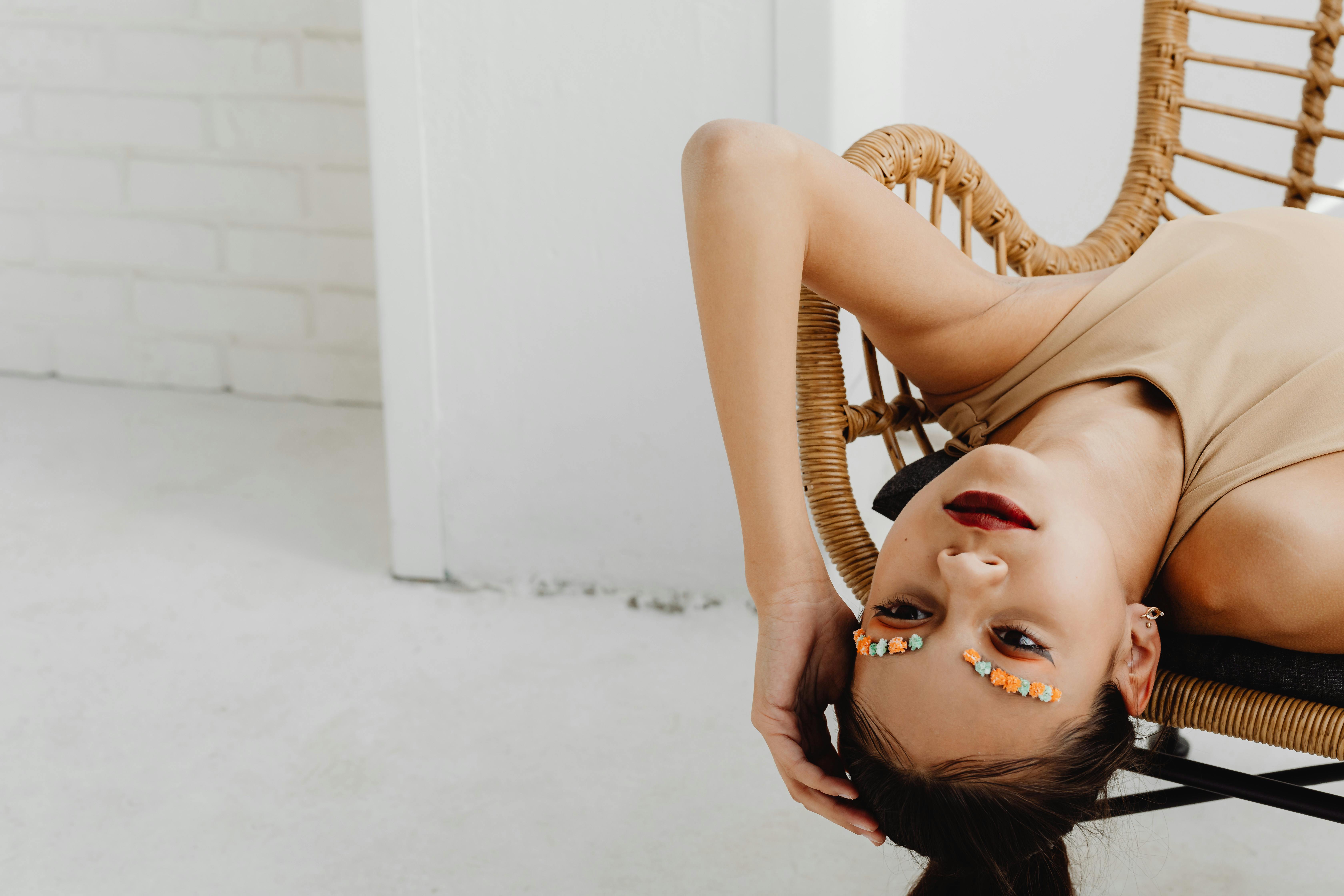 Upside-down portrait of a woman with floral eyebrows and red lips on wicker chair.