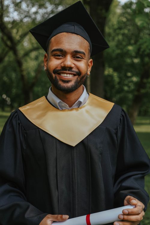 Free Young adult graduate smiling in cap and gown, celebrating achievement outdoors with diploma. Stock Photo