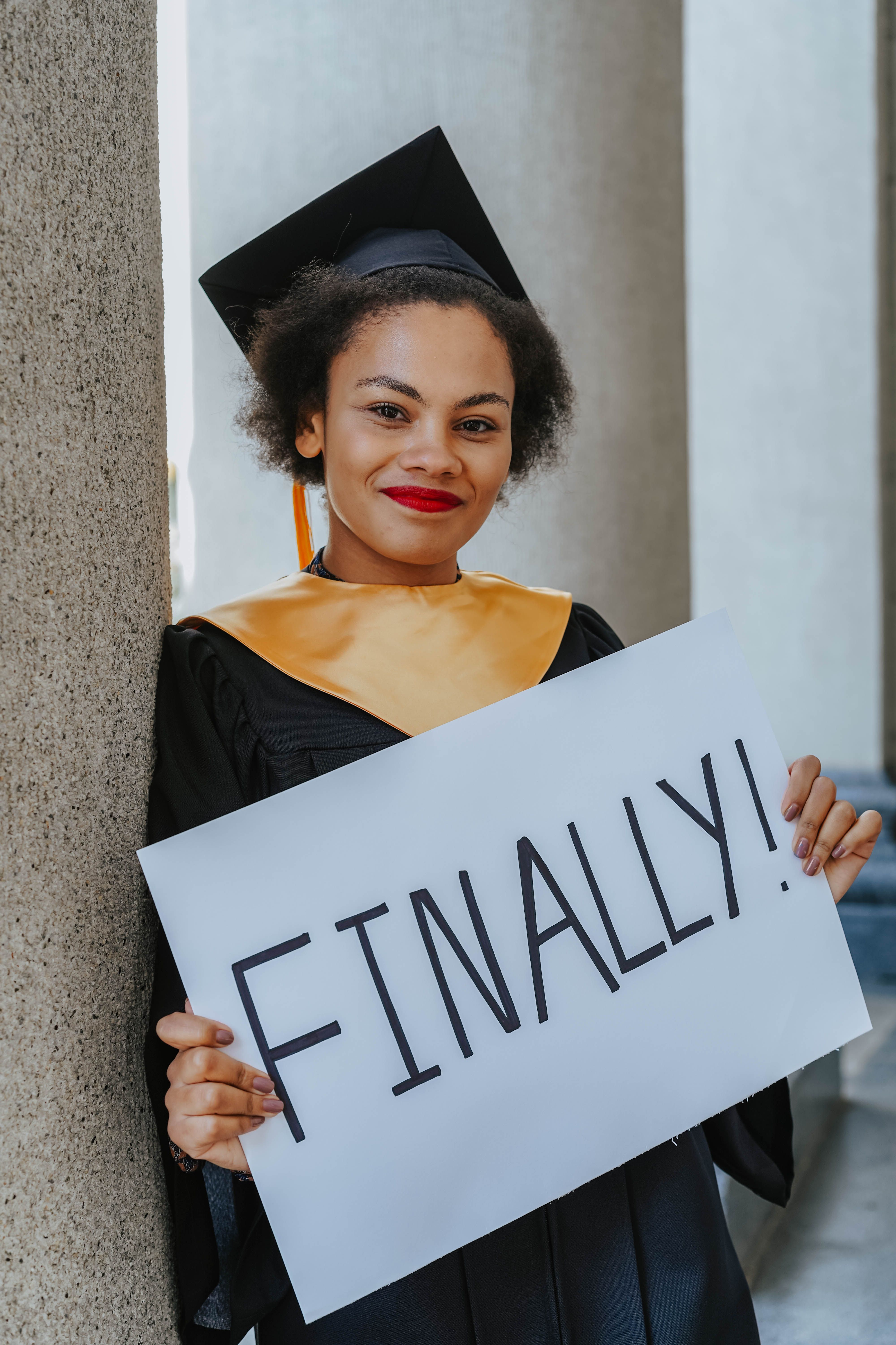 Young woman in graduation gown holding a 'Finally!' sign, celebrating her achievement.