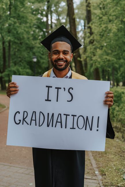 A joyful graduate proudly holds a sign saying 'It's Graduation!' in a lush outdoor setting. A joyful graduate proudly holds a sign saying 'It's Graduation!' in a lush outdoor setting.