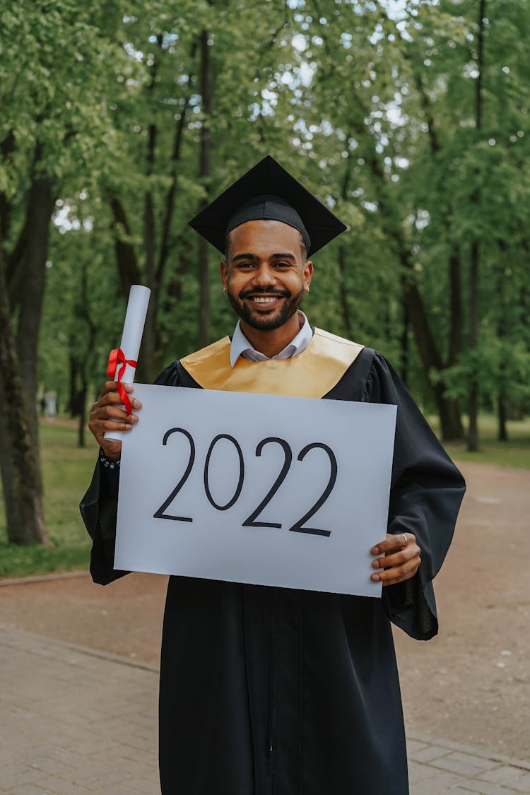 A Man In Black Toga Holding A Signboard And Diploma