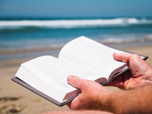 Free Hands holding an open book at a beach with waves in the background, perfect for summer relaxation. Stock Photo