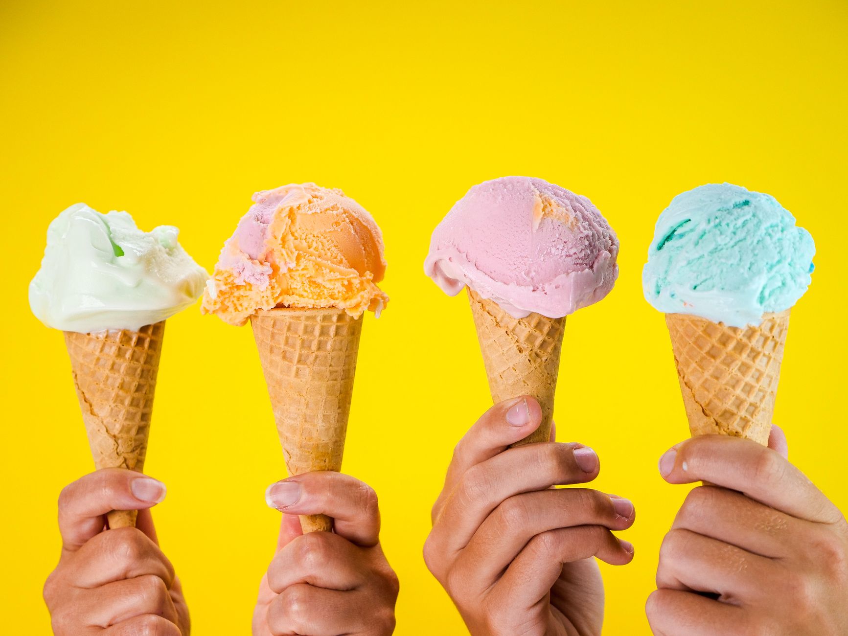 Four colorful ice cream cones held up against a bright yellow backdrop, showcasing various flavors.