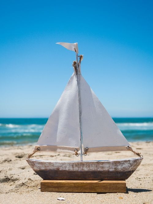 Free A charming wooden toy sailboat on a sunny beach with clear blue skies and ocean waves. Stock Photo