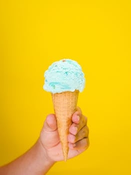 Close-up of a blue ice cream cone held by a hand against a vivid yellow background.