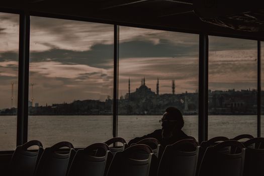 A silhouette of a man sitting on a ferry as the Istanbul skyline and Hagia Sophia are visible at dusk.