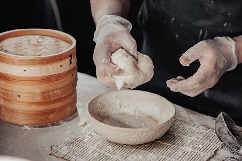 Free Close-up of hands kneading dough beside a bamboo steamer in a kitchen setting. Stock Photo