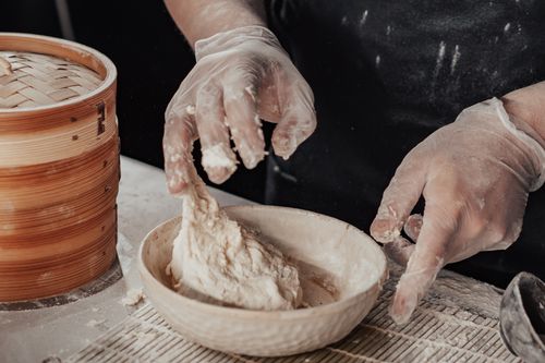 Free Close-up of hands wearing gloves kneading dough in a ceramic bowl beside a bamboo steamer. Stock Photo