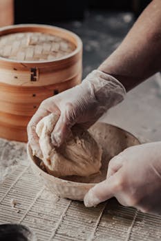 Close-up of hands kneading dough in a rustic kitchen setting with bamboo steamer visible.