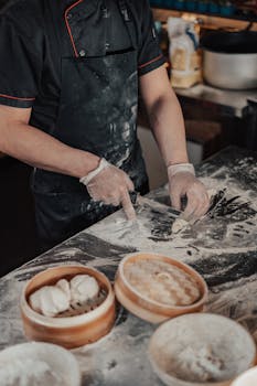 Chef's hands making dumplings from dough on a floured surface indoors.