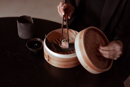 Free A close-up of a person using chopsticks to pick up a dumpling from a bamboo steamer on a dark table. Stock Photo