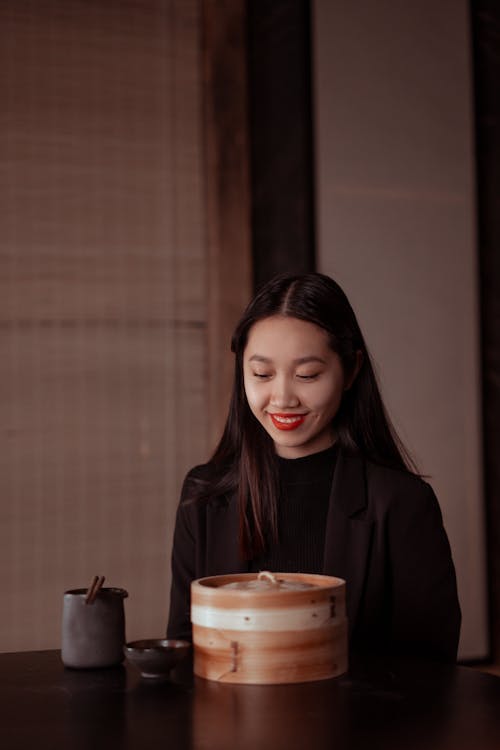 Smiling Young Woman Sitting by Bamboo Steamer on Table