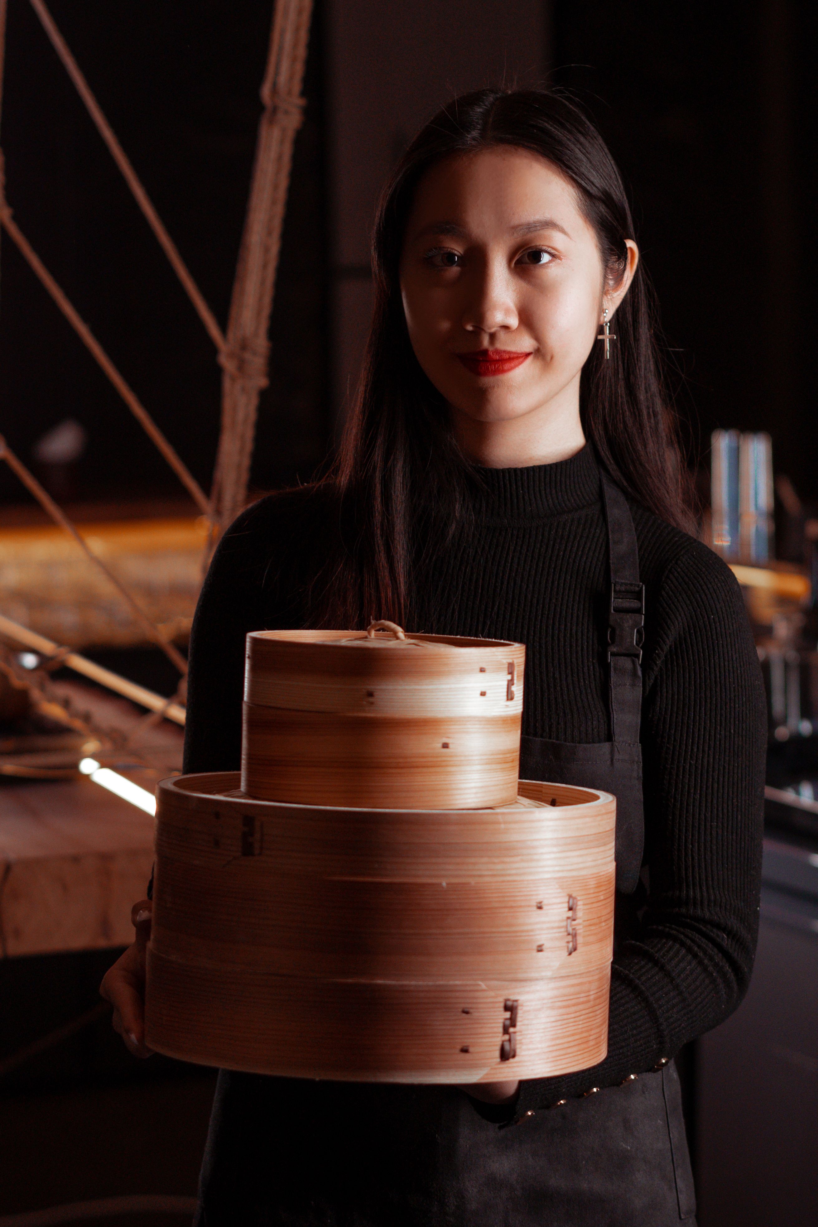 A stylish woman holds bamboo steamers in a dimly lit restaurant, exuding warmth and charm.