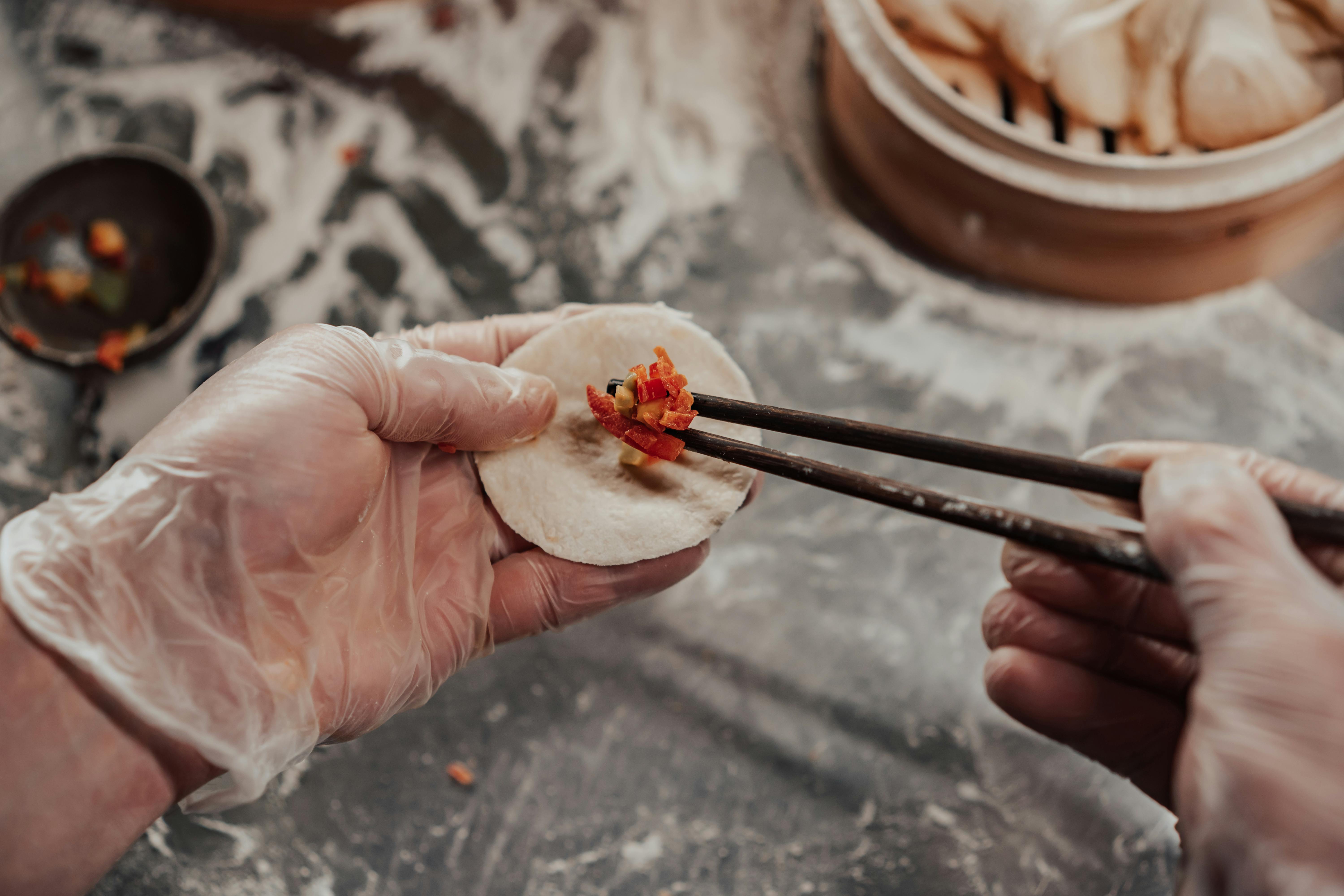 Close-up of gloved hands using chopsticks to stuff dumplings in a kitchen setting.