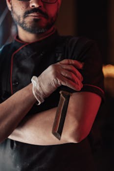 A professional chef holding a knife, wearing gloves and a dark uniform in a dimly lit kitchen.