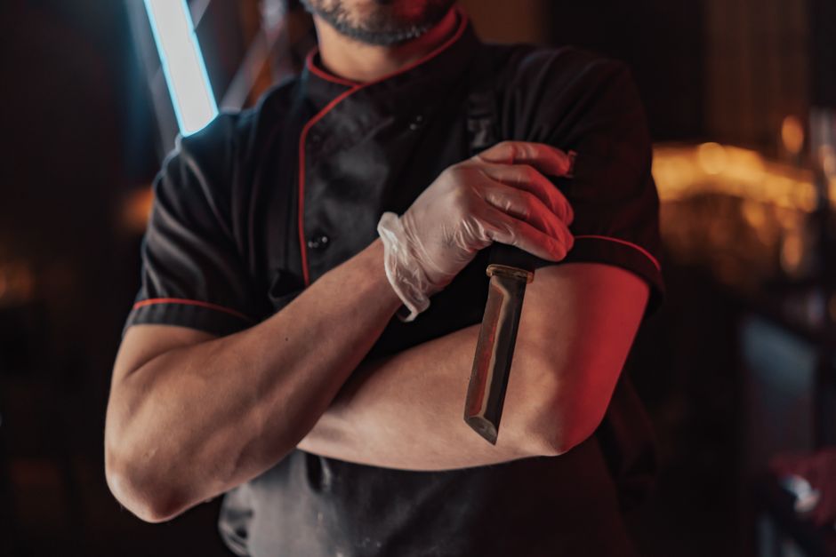Chef in black uniform with knife and latex gloves in a dimly lit kitchen setting.