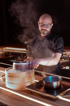 Professional chef in a kitchen using a bamboo steamer to prepare food, surrounded by steam.