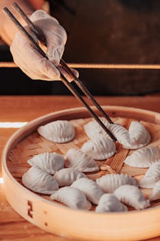 A chef's hand arranging dumplings in a bamboo steamer with chopsticks.