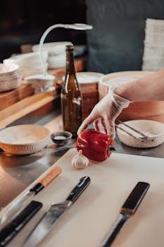Gloved hand holding red bell pepper on chopping board with various kitchen tools.