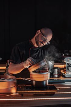 A chef in a black uniform prepares dim sum using a bamboo steamer in a professional kitchen.