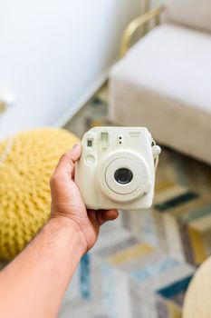 Close-up of a hand holding a white Instax camera in a modern indoor setting.