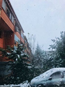 A snowfall over an apartment building and car in a wintery Istanbul street.