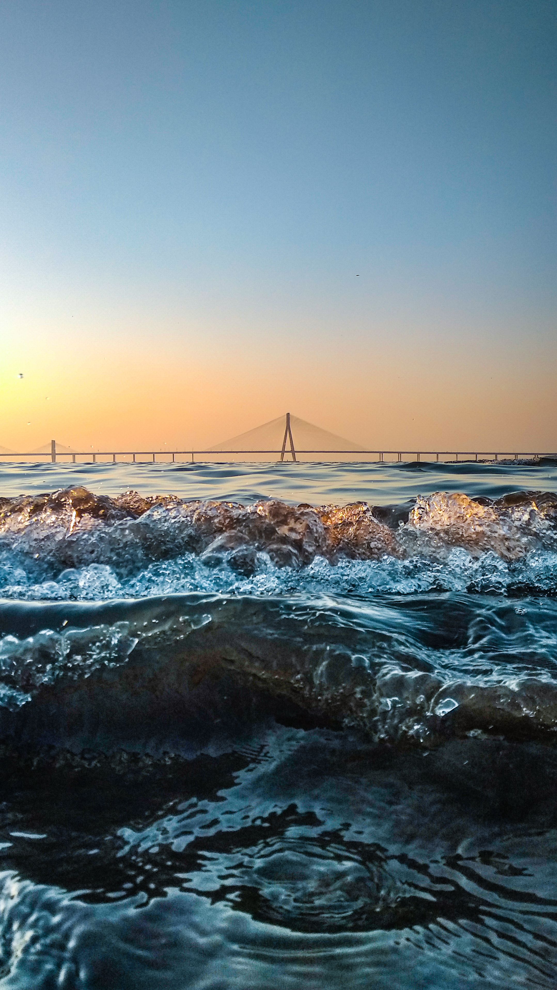 Dramatic ocean waves crashing at sunset with a beautiful bridge in the distance.