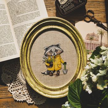 A beautifully framed raccoon cross stitch with gardening theme, surrounded by open book and flowers.（イメージ）