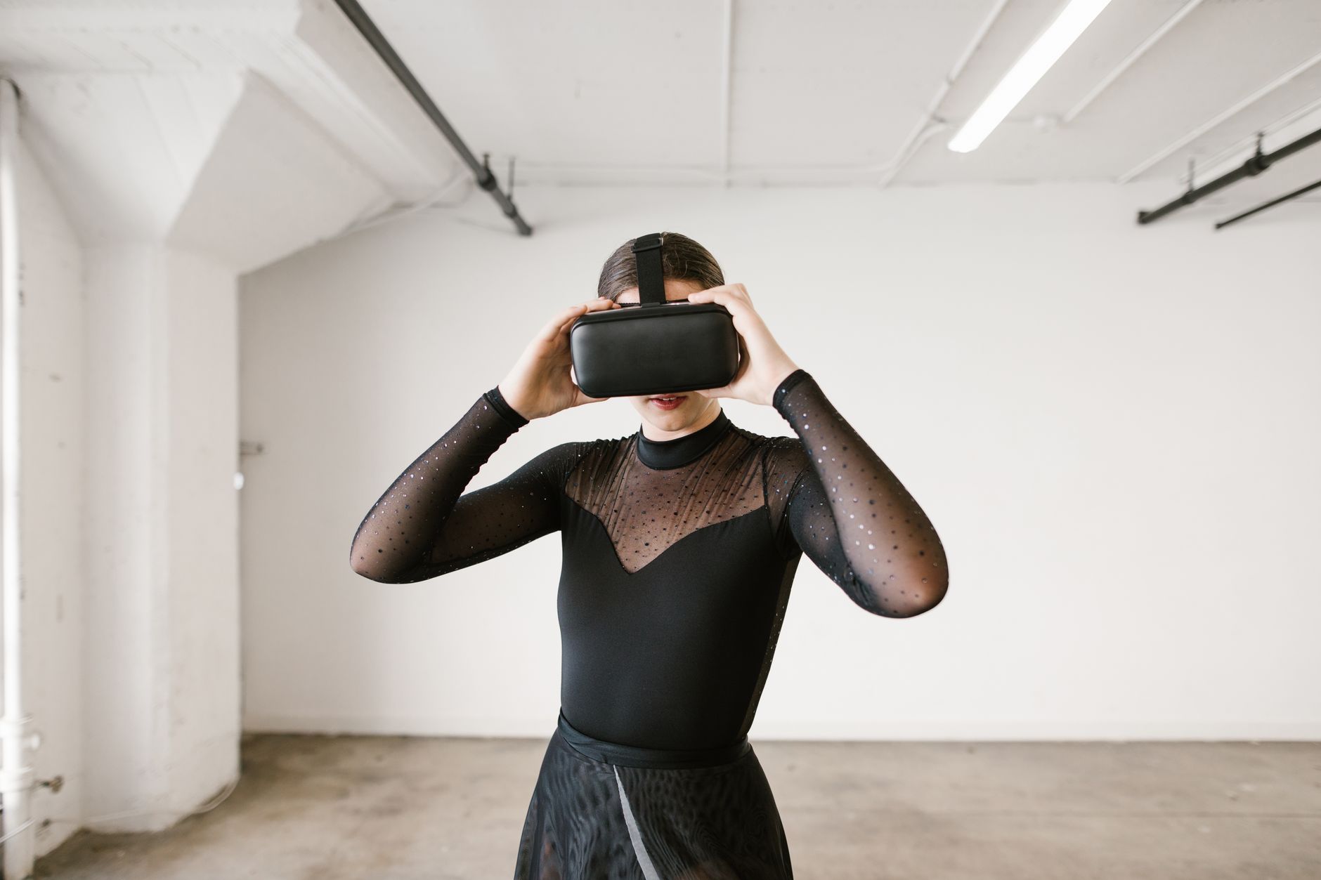 A ballerina in black practicing ballet with a VR headset in a minimalistic indoor setting.