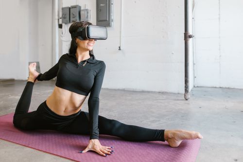 Free Smiling woman in black yoga attire stretches with virtual reality goggles on a mat indoors. Stock Photo
