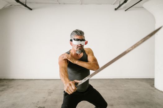 Adult man wearing VR headset practicing sword techniques in empty indoor space.