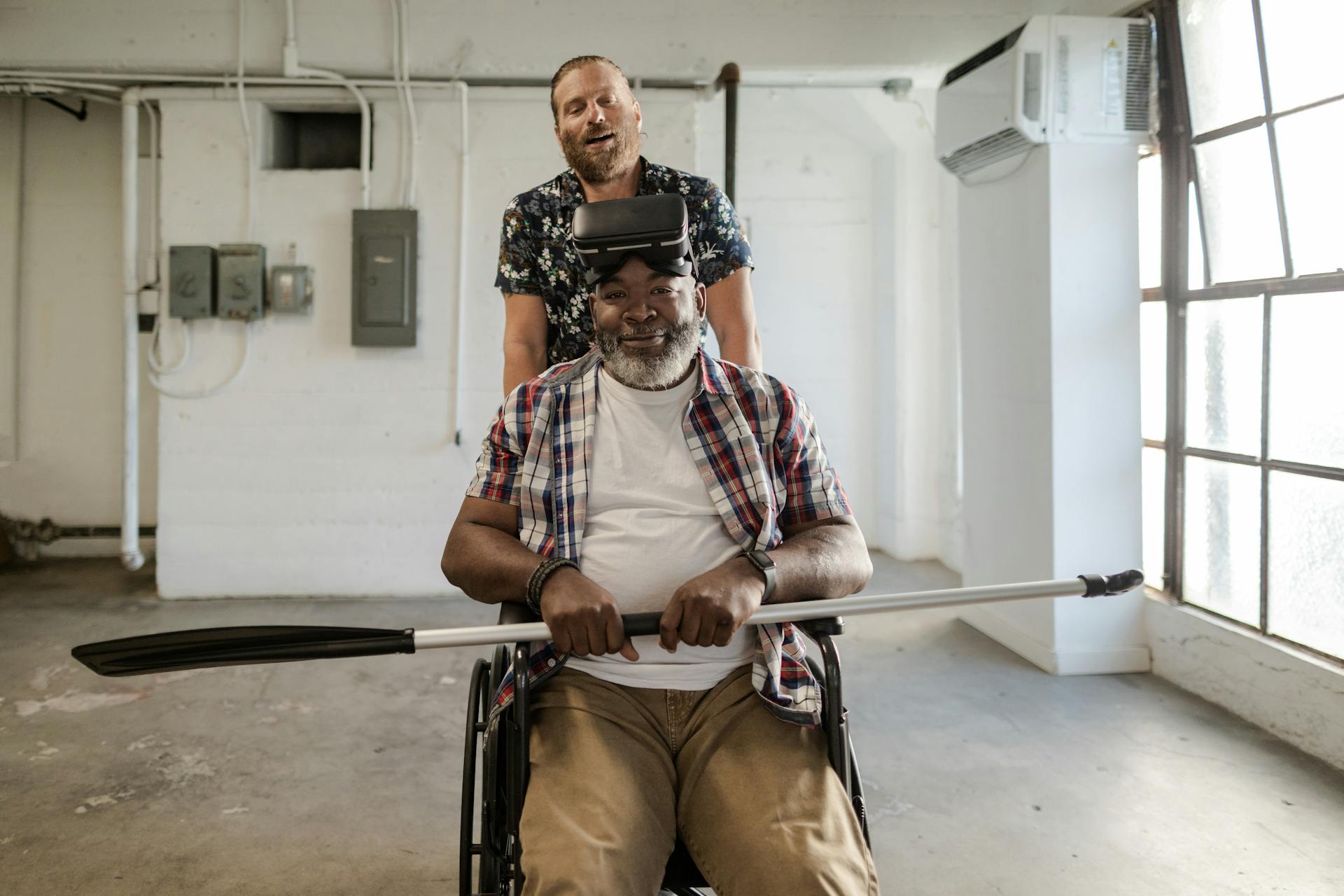 Two men enjoy virtual reality therapy indoors, one in a wheelchair holding a paddle.