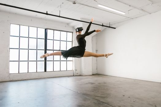 Ballet dancer wearing VR headset performs a leap in sunny studio, blending classical dance with modern technology.